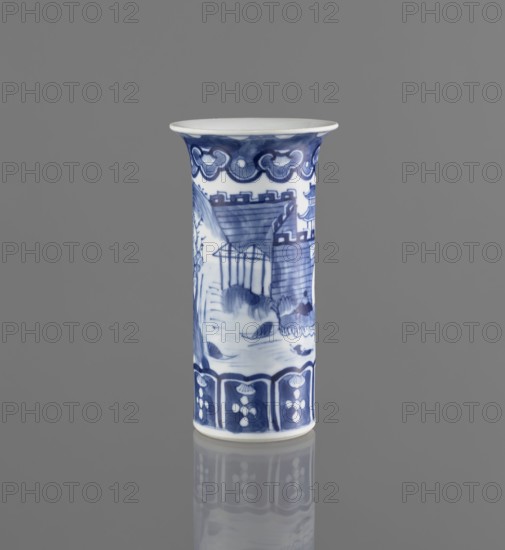 Flute-type vase, ceramics, blue-and-white porcelain, Chinese art, landscape motifs, Chinese, 17th/18th century, China, vessel, height 15 cm, diameter 8.1 cm, A tall cylindrical ceramic vase stands upright against a solid gray background, painted in white with blue decorative motifs; the rim flares slightly outward and is white on top with a blue border beneath, a band of stylized cloud-like blue patterns encircles the upper section, while the main body features a blue landscape scene with architectural shapes, sloped rooftops, leafy elements, and rounded forms, and the lower section shows a repeating blue panel design with floral-like white shapes; the vase sits on a reflective surface that shows a muted mirror image of its base and lower decorations.