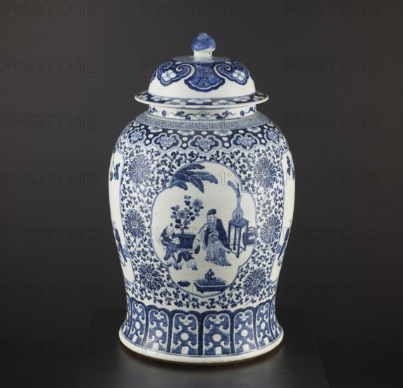 Pot with cover, blue-and-white porcelain, Chinese; plant motifs and mountain landscapes; decorative vessel; ceramics in Qing style; possibly 1st half of the 17th century; China; height 60 cm, diameter 36 cm; vessel; Qing 1644–1911 and Ming 1368–1644 periods noted; repossession provenance., A lidded ceramic jar decorated in blue and white sits against a dark gray background; the jar has a rounded body with a flared base and a domed lid topped by a small knob, all covered in intricate blue painted patterns on a white ground, including floral and scrolling motifs around the neck and body, a framed pictorial scene on the front showing human figures near a small table and plant, stylized floral medallions and foliate scrollwork filling the surrounding areas, a decorative band of repeating geometric and curving elements near the base, and blue detailing on the lid and rim.