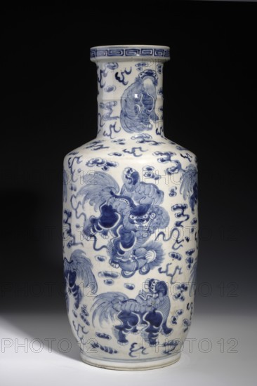 Tall bang chui vase, Chinese, Qing dynasty Kangxi period, 1662–1722, 17th century. Blue-and-white porcelain vessel, height 46.0 cm. Wide cylindrical neck terminating in a round ring, angular shoulders and an oval belly gently tapering toward the bottom, overall shape resembling a wooden hammer/pestle. Surface densely painted with fo lions playing with a rag ball among clouds; ring around the neck decorated with a meander., A tall ceramic bottle-shaped vase with a narrow neck and flared rim sits against a dark background, its surface predominantly white with painted blue decorative motifs; the blue designs include stylized cloudlike swirls and several mythical lionlike creatures in dynamic poses distributed across the body and neck, with detailed fur, expressive faces, flowing manes, and curled tails; a band of geometric blue pattern encircles the rim, and the base shows a thin blue line; the overall visible colors are white and shades of blue against a black and gray backdrop.