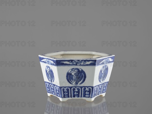Flowerpot, Chinese, Qing dynasty 1644–1911, blue-and-white porcelain jardinnière decorated with lotuses, flowers, birds and phoenixes, 18th–19th century, China; vessel, height 15.2 cm, A hexagonal ceramic container sits on a reflective surface against a plain gray background, showing a white base color with blue decorative patterns; each side features a circular blue medallion with a bird-like figure, a blue band with floral and cloud-like motifs near the rim, and a lower blue frieze with repeating vertical decorative panels, while the interior appears pale and the underside shows small unglazed patches.