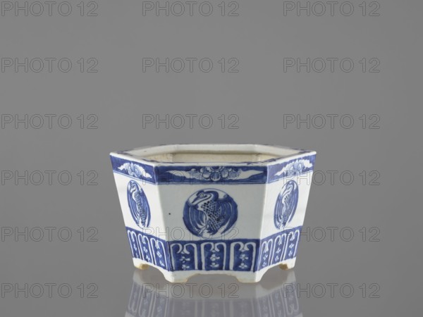 Flowerpot, Chinese, Qing 1644–1911, 18th/19th century. Blue-and-white porcelain jardinnière with floral decorations—lotuses and flowers—and birds including phoenixes. Vessel; height 15.2 cm., A hexagonal ceramic vessel with an open top, shown against a neutral gray background, sits on a reflective surface. The vessel is predominantly white with cobalt blue painted decoration, including a band of floral and cloud-like motifs near the rim and a lower band of repeating vertical panels with stylized floral elements. Each face features a circular medallion with a bird motif rendered in blue. The vessel stands on short bracket feet, and the interior appears unglazed or lightly stained compared with the exterior. The visible colors are white, cobalt blue, gray, and a small area of beige on the feet.