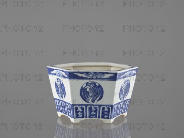Flowerpot, Chinese, Qing dynasty 1644–1911, 18th–19th century. Blue-and-white porcelain jardinière decorated with floral motifs—lotuses and other flowers—and birds including phoenixes. Vessel, height 15.2 cm., A hexagonal ceramic container with a glossy white base and blue painted decoration sits on a reflective surface against a gray background; each side features a circular blue medallion with a bird motif and a patterned blue band near the top and bottom, and the piece rests on short feet with visible wear at the edges.