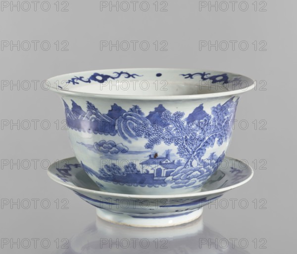 Flowerpot on base, Chinese, Qing dynasty 1644–1911, blue-and-white porcelain jardinière with landscape motifs and floral decorations, flowers, 18th–19th century, China; vessel, height 19.7 cm, diameter 28.2 cm., A ceramic bowl with a matching saucer, both decorated in blue on a white ground; the bowl has a flared rim and a rounded body sitting on a short foot, and the saucer mirrors the bowl's curved profile. The painted decoration shows a landscape scene wrapping around the bowl with a group of trees, a small building near the water, stylized cloud and wave motifs, and distant mountain shapes, while the rim and saucer edge are accented with ornamental scroll-like motifs. Visible colors include shades of blue and white, with a small area of brownish discoloration.