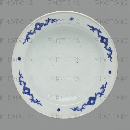 Flowerpot on base, Chinese, Qing 1644–1911, blue-and-white porcelain jardinière with landscape motifs and floral decorations, flowers; 18th–19th century, China; vessel; height 19.7 cm, diameter 28.2 cm., A round ceramic plate with a pale off-white surface and a glossy finish, decorated around its rim with a few stylized floral motifs and curving vine-like patterns in a deep blue, plus a few small solid blue dots spaced along the inner rim; the background behind the plate is a flat medium gray.