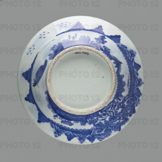 Flowerpot on base;, Chinese; Qing dynasty 1644–1911; blue-and-white porcelain jardinière with landscape motifs and floral decorations; 18th–19th century; vessel; height 19.7 cm, diameter 28.2 cm., A round ceramic plate shown from above with a central recessed foot and a wide rim decorated in blue on a white ground; the decoration includes a landscape scene with stylized mountains, trees, and water rendered in varying shades of blue, a few small bird-like shapes, a boat-like form, and ornate patterned borders; the foot rim shows a worn beige edge and the plate surface has small dark specks; visible colors include blue, white, beige, gray, and a hint of brown.