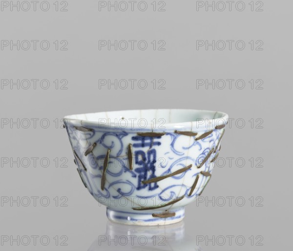 Bowl, gift, Chinese, Qing dynasty 1644–1911, 19th century; blue-and-white porcelain bowl decorated with flowers; ceramic vessel; height 5.8 cm, diameter 9 cm; conservation: clasps., A small porcelain bowl with a rounded rim and a short foot, predominantly white with blue painted swirling patterns and decorative motifs, shows a group of metallic staples or repairs affixed across its surface and visible hairline cracks in the glaze; the bowl sits on a reflective surface against a neutral gray background, and the visible colors are white, blue, metallic brownish-gold, and gray.