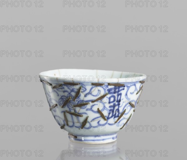 Bowl, gift; Chinese, Qing 1644–1911, 19th century; blue-and-white porcelain vessel decorated with flowers; height 5.8 cm, diameter 9 cm; conservation techniques: clasps., A small ceramic cup with a white base and blue painted floral and scroll patterns around its exterior, showing a group of metallic staples or pins holding visible cracks together; the cup has a flared rim, a narrow foot, and glaze reflections, with colors including white, blue, brownish metal, and hints of green against a neutral gray background.