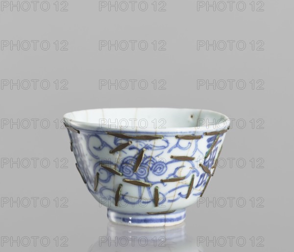 Bowl, Chinese, Qing 1644–1911, 19th century, blue-and-white porcelain vessel decorated with flowers, conservation techniques — clasps; height 5.8 cm, diameter 9 cm., A small ceramic bowl with a rounded shape and a narrow foot, predominantly white with blue decorative brushwork forming swirling and floral patterns on its exterior; the surface shows visible cracks repaired with metal staples that are brass or bronze in color, some staples showing a slight greenish patina; the rim and foot have thin blue bands, and the interior is mostly white with faint hairline cracks; the background is a plain light gray.