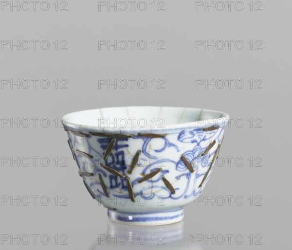 Bowl, Chinese, Qing dynasty 1644–1911, 19th century. Blue-and-white porcelain vessel decorated with flowers. Height 5.8 cm; diameter 9 cm. Conservation: clasps., A small ceramic bowl with a rounded shape and a narrow foot, showing a white base color with blue decorative brushwork forming floral and scrolling patterns, and a few metal staples or strips affixed across visible cracks and joins; the rim and foot have subtle blue bands, and the interior appears pale with faint hairline lines; the visible colors are white, blue, metallic brown, and hints of green against a neutral gray background.