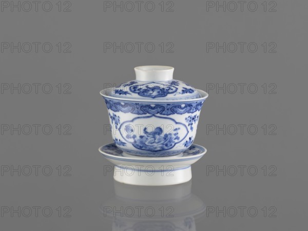 Bowl on a base with a lid, ceramics, blue and white porcelain, Chinese art, female images, figural decorations, Chinese, 19th century, South China, China, vessel, height 8.6 cm, A white porcelain lidded cup and saucer with blue decorative patterns sits centered on a reflective grey surface against a grey background; the cup has ornate blue floral and scrolling motifs around its body and lid, a flared rim, and a short neck, while the saucer and the raised base show matching blue border patterns.