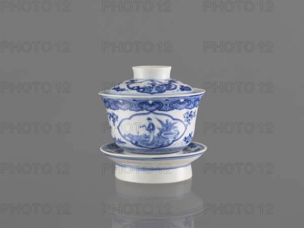 Bowl on a base with a lid, blue-and-white porcelain, Chinese, 19th century, South China, vessel, height 8.6 cm — figural decorations depicting female images., A white porcelain cup with a lid and matching saucer decorated with intricate blue painted floral and figurative patterns sits centered against a plain gray background, showing a smooth glossy surface and a subtle reflection beneath; the colors visible are white, various shades of blue, and gray.