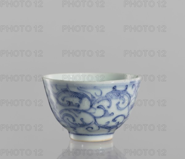 Bowl, blue-and-white porcelain, ceramics; floral ornaments depicting flowers; 19th century, China; vessel; height 4.3 cm, diameter 6.7 cm; ., A small ceramic cup with a rounded shape sits on a reflective surface against a plain gray background; the cup is primarily white with intricate blue swirling patterns covering its exterior, a pale blue interior, a thin darker blue band near the base, and a narrow unglazed rim at the bottom showing a beige tint, with glossy highlights on the glaze.