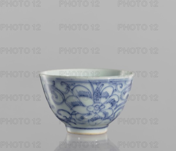 Bowl, blue-and-white porcelain, ceramics, Chinese, 19th century, floral ornamentation, vessel, height 4.3 cm, diameter 6.7 cm, A small ceramic cup sits against a plain light gray background, showing a pale off-white interior and exterior decorated with delicate blue floral and scroll patterns; the cup has a gently flared rim, a rounded body tapering to a short foot with a thin blue band above the base, and a glossy finish that produces subtle highlights and a soft reflection on the surface beneath it, with visible colors including off-white, several shades of blue, light gray, and a hint of pale tan at the foot edge.