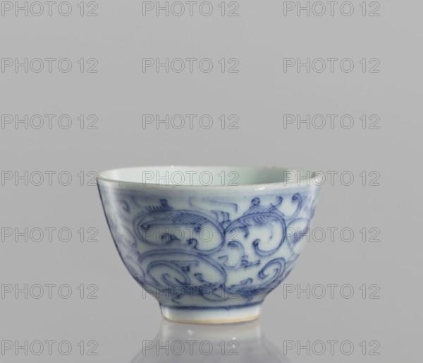 Bowl, flowers, blue and white porcelain, ceramics, Chinese, floral ornaments, 19th century, China, vessel, height 4.3 cm, diameter 6.7 cm, A small ceramic bowl sits on a reflective surface against a plain background, showing a smooth rounded shape with a slightly flared rim and a narrow foot. The bowl is primarily white with an overall glossy finish and features a flowing hand-painted pattern in blue that wraps around its exterior, consisting of curved, swirling motifs and fine brushwork. The interior appears plain and pale, and the bottom edge of the foot has a subtle warm off-white to light beige tone. The visible colors in the image include shades of white, blue, light beige, and gray.