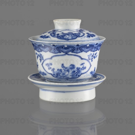 Bowl on a base with a lid;, Ceramic, blue-and-white porcelain, 19th century, South China, China. Figural decorations depicting female images. Vessel; height 8.6 cm., A porcelain lidded cup and saucer set sits against a gray background, the pieces being white with blue hand-painted decorative motifs. The cup has a flared rim and a domed lid with a small necked finial, both showing intricate blue floral and cloud-like patterns and a central scene on the cup body featuring a small bird among foliage. The saucer mirrors the blue border motifs and supports the cup on a short white foot, and a soft reflection of the set appears on the surface below. Colors visible include white, blue, and gray.