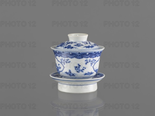 Bowl on a base with a lid, ceramic blue-and-white porcelain, Chinese, figural decorations depicting female images, 19th century, South China, China, vessel, height 8.6 cm, A small lidded ceramic cup with a matching saucer sits centered on a reflective surface, both showing a white base color with intricate blue floral and decorative patterns; the lid has a small raised knob and blue motifs, the cup flares outward slightly at the rim with ornate blue borders and floral clusters, and the saucer echoes the white and blue design with concentric blue lines; the background and reflection are shades of gray.