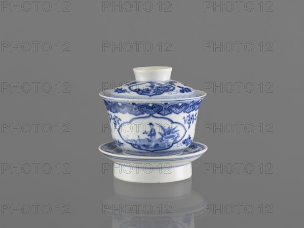 Bowl on a base with a lid, ceramics, blue and white porcelain, Chinese art, figural decoration depicting female images, 19th century, South China, China, vessel, height 8.6 cm, A white porcelain cup with a lid and saucer is shown against a solid gray background, the cup and lid decorated with blue painted floral and ornamental motifs and rounded cartouche shapes, with a blue border near the rim; the saucer and the cup's foot also have blue rim accents, and the glossy surface of the porcelain produces subtle highlights and a soft reflection on the gray surface beneath.