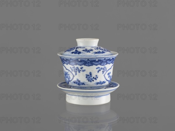 Bowl on a base with a lid, ceramics, blue and white porcelain, Chinese art, female images, figural decorations, 19th century, South China, China, vessel, height 8.6 cm, A white porcelain cup with a lid and a matching saucer sits centered against a gray background, each piece decorated with blue painted floral and ornamental patterns; the cup has a flared rim and rounded body with blue motifs around the upper and lower sections, the lid has a small raised knob and blue floral designs, and the saucer has a blue rim and echoed decorative band, all reflected faintly on the glossy surface beneath.