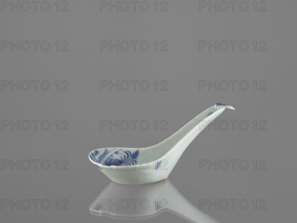 Sauce boat spoon, Chinese, blue and white porcelain with plant ornaments, tableware element vessel, Qing dynasty, mid-19th century, China; length 19.2 cm., A small ceramic spoon with a long handle sits on a reflective surface against a plain gray background, the spoon being pale off-white with blue painted floral motifs visible on the inside bowl and small blue spots along the handle, and its reflection visible below showing similar off-white and blue tones.