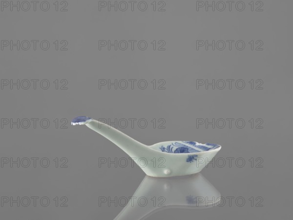 Sauce boat spoon, Chinese, blue-and-white porcelain with plant ornaments, tableware element vessel, Qing dynasty, mid-19th century, China, length 19.2 cm., A small ceramic spoon with a long slender handle and a shallow bowl sits on a reflective surface, showing a faint mirror image below; the spoon is primarily pale white with blue painted decoration visible inside the bowl and on the tip of the handle, and the overall scene has a neutral gray background.