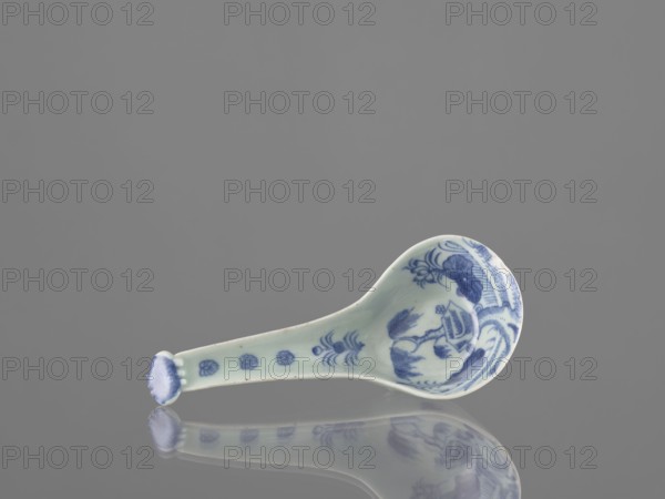 Sauce boat spoon, Chinese, blue-and-white porcelain with plant ornaments, tableware element, ceramic vessel, Qing dynasty mid-19th century, China; length 19.2 cm., A small ceramic spoon lies on a reflective surface, showing a mirrored reflection beneath it. The spoon is pale with blue decorative patterns concentrated on the bowl and along the handle, including floral and abstract motifs. The background is a uniform gray, and the reflection shows the same pale and blue colors reversed on the surface.