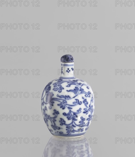 Snuffbox with spoon, Chinese, 19th century, blue and white porcelain, plant ornaments and floral tendril decoration, height 6.8 cm, vessel., A small rounded ceramic bottle with a short neck and a domed stopper sits upright on a reflective surface, decorated with an intricate floral and vine pattern in blue on a white background; the stopper is darker blue and the neck has a narrow blue band, while the reflected image shows softened blues and whites against a light gray backdrop.