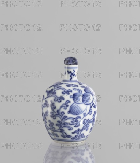 Snuffbox with spoon;, ; Chinese; 19th century; blue-and-white porcelain; plant ornaments with flowers and floral tendril; vessel; height 6.8 cm., A small rounded ceramic bottle with a narrow neck and a rounded stopper sits centered against a plain light gray background, the bottle showing a white base color with blue intricate floral and leaf patterns wrapping around its body and neck, and the stopper appearing dark blue to indigo with a marbled texture; the bottle rests on a reflective surface that creates a faint mirrored image beneath it.