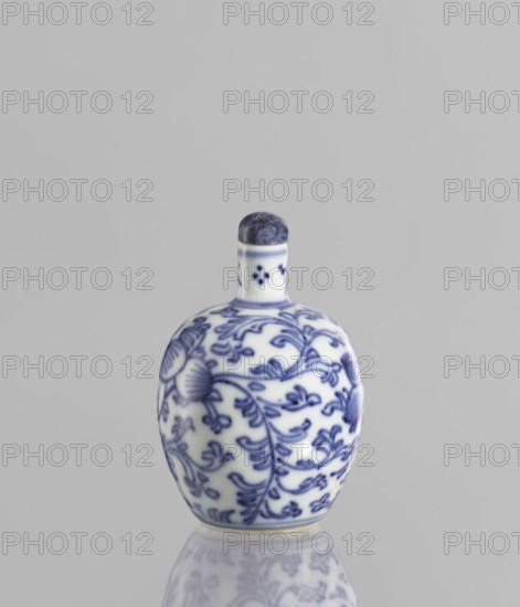 Snuffbox with spoon, blue-and-white porcelain, 19th century China; plant ornaments and floral tendril decoration; vessel; height 6.8 cm., A small rounded ceramic bottle with a short neck and a rounded stopper sits against a plain light gray background, the bottle showing a white base color with a blue floral vine pattern covering its surface and a darker blue mottled stopper, the ceramic surface reflecting faintly below to create a subtle mirrored image; visible colors include white, several shades of blue, gray, and a hint of off-white at the bottle's base.