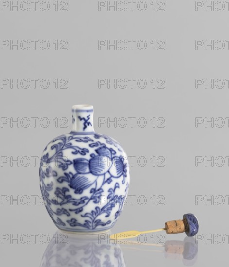 Snuffbox with spoon, blue-and-white porcelain, China, 19th century; plant ornaments with flowers and floral tendril; vessel, height 6.8 cm., A small porcelain bottle with a short neck sits on a reflective surface, decorated with blue floral and vine patterns on a white background; beside it lies a cork with a blue-topped stopper attached to a pale yellow spoon-like implement, and the overall colors visible are white, blue, pale yellow, tan, and gray.