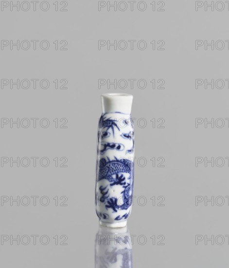 Flask - snuffbox, Chinese, blue and white porcelain, ceramics; pearls, clouds, dragons iconography; Kangxi style; Qing 1644–1911, 19th century; vessel; height 7 cm; ., A small cylindrical ceramic object stands upright against a plain light gray background, predominantly white with intricate blue painted designs wrapping around its surface; the blue motifs include a stylized dragon and flowing decorative patterns. The top is slightly flared and white, and the base tapers gently with a glossy sheen that produces a subtle reflection on the smooth surface beneath, where a faint mirrored image of the object appears. The visible colors are white, blue, light gray, and a hint of darker gray in the reflection.