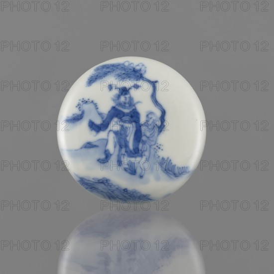 Seal ink box, depiction of Kui Xing iconography, ceramics, Chinese blue and white porcelain, writing implement, Qing dynasty 1644–1911, 19th century, China; vessel; height 2 cm, diameter 6.2 cm., A round porcelain disk with a glossy white surface decorated in blue, showing a painted scene of a figure wearing flowing garments beneath a curved tree branch, with another smaller figure nearby, stylized rocks and foliage at the base, and a soft reflection of the disk visible on the gray background; colors visible include white, blue, and gray.