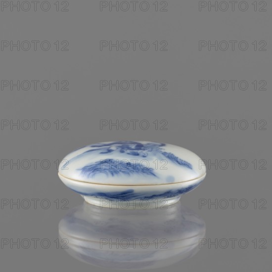 Seal ink box, Kui Xing iconography, ceramics, blue and white porcelain, writing implement, Qing dynasty 1644–1911, 19th century, China; vessel, height 2 cm, diameter 6.2 cm., A small oval ceramic box with a domed lid sits on a reflective surface, showing a soft reflection beneath it; the box is predominantly white with flowing blue painted designs resembling stylized waves or foliage, and a thin band of pale gold along the lid's edge; the background is a uniform mid-gray.