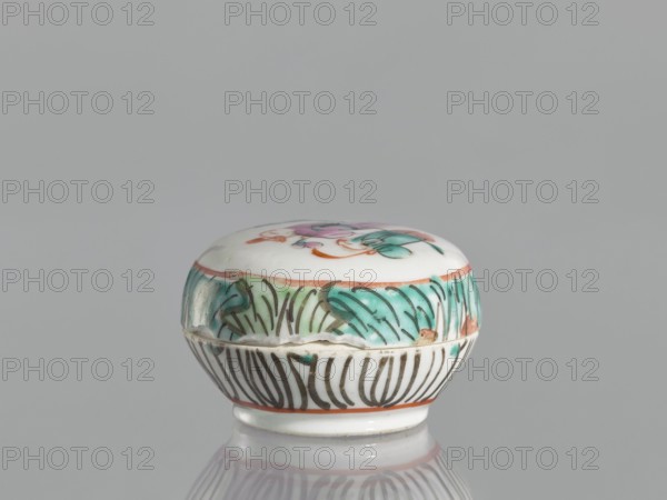 Box with a lid, Chinese, colored porcelain with figural scenes, 19th century, vessel, height 4.6 cm, width 6.9 cm, A small round lidded porcelain container sits on a reflective surface against a neutral gray background, showing a white base color with painted decorations in green, turquoise, pink, orange, brown, and black; the lid top has floral motifs in pink and green with orange accents, the upper rim band displays a green and turquoise leafy pattern with brown outlining, and the lower body is decorated with vertical black stylized lines and an orange band near the foot.
