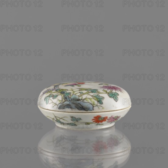 Lidded box, Chinese, colored porcelain decorated with pomegranates, chrysanthemums and peonies, floral ornaments; mid-19th century Qing dynasty, 1644–1911; ceramic vessel for writing instruments; height 3.2 cm, diameter 7.9 cm., A small oval porcelain box with a domed lid sits against a plain gray background, its surface predominantly white with painted botanical motifs in green, blue, purple, red, pink, and orange, showing leaves, flowers, and a bluish rock-like shape; the box has a thin gold-colored rim where lid meets base and rests on a reflective gray surface that creates a soft mirrored image of the painted floral decoration.