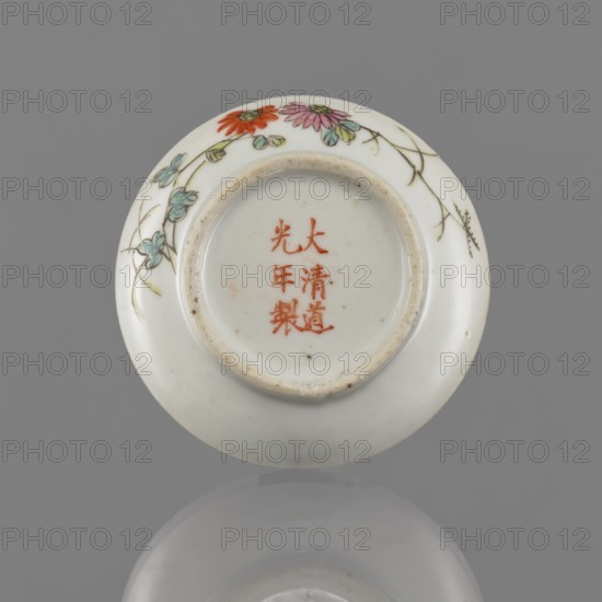 Lidded box, Chinese, colored porcelain decorated with pomegranates, chrysanthemums and peonies—floral ornaments; ceramic vessel used for writing instruments; Qing dynasty mid-19th century, 1644–1911; height 3.2 cm, diameter 7.9 cm., A round white ceramic object with a recessed base is shown flat against a gray background, decorated near the rim with painted flowers and trailing stems in colors including red, pink, green, turquoise, and pale yellow, with fine brown outline lines; the central recessed area shows a few small brown specks and the rim of the recess has a slightly rough, beige-tinged edge; the surface reflects softly onto the gray background creating a muted mirror image.
