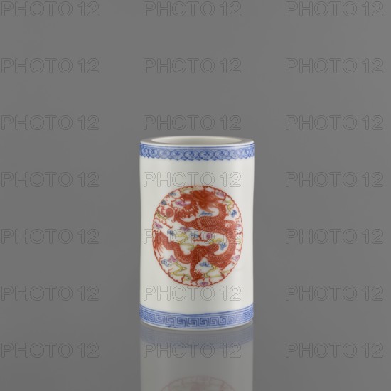 Brush vessel, Chinese, 19th century, ceramics; decorated with dragons and meander ornament; function: vessel for brushes; height 11.4 cm, diameter 7.1 cm; ., A cylindrical white ceramic vessel with decorative blue borders at the top and bottom, featuring a circular red dragon motif surrounded by small colorful cloud-like shapes on the front; the background is a smooth gray, and the vessel casts a subtle reflection on the surface below.