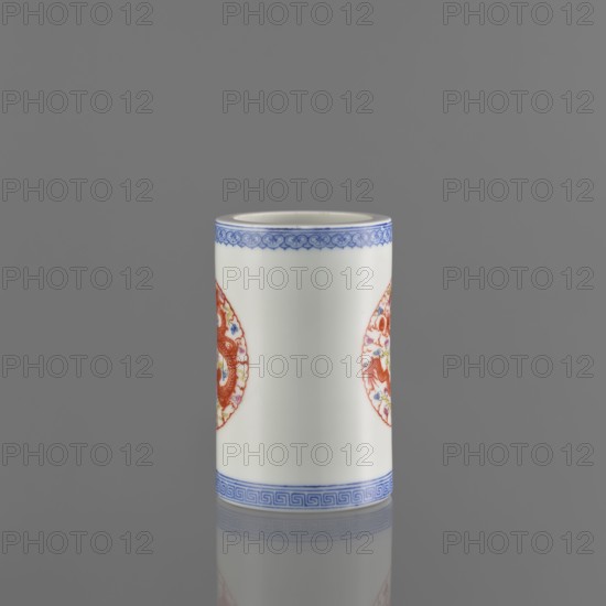 Brush vessel, Chinese, 19th century, ceramics, decorated with dragons and meander ornament, function: vessel brush holder, height 11.4 cm, diameter 7.1 cm., A cylindrical white ceramic vessel with decorative blue bands near the top and bottom edges and a pair of circular red ornamental medallions on its sides, shown standing on a reflective gray surface against a plain gray background. The visible colors include white, blue, red, and gray.