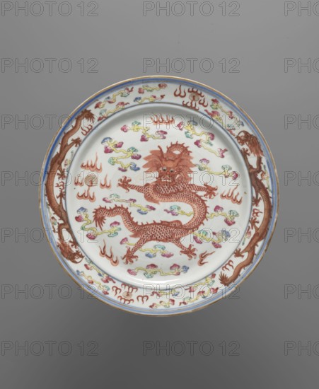 Plate, Chinese, Qing dynasty Guangxu, 1875–1908, late 19th century. Colored porcelain vessel decorated with dragons, clouds, and flames. Height 2.6 cm; diameter 25.8 cm., A round ceramic plate with a white background featuring a central red dragon with detailed scales and an expressive face, surrounded by a few cloud-like motifs in colors including yellow, green, blue, and pink; the rim has repeating red dragon figures and similar multicolored cloud elements, with a thin blue band near the edge and a narrow gold outer edge; the overall visible colors include white, red, blue, yellow, green, pink, and gold, set against a gray background.