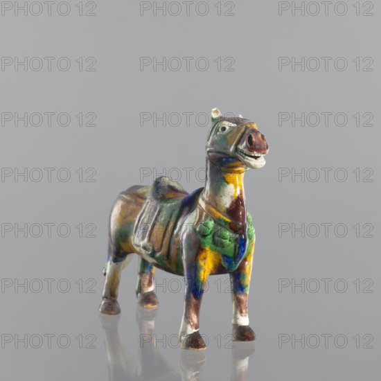 Mule, Chinese, Qing dynasty 1644–1911, 18th century. Colored porcelain animal figurine sculpture of a mule. Height 19.1 cm, width 19.4 cm., A small ceramic figurine of a horse stands on a reflective surface against a plain gray background, the figure glazed in a mix of green, yellow, blue, brown, and white; the horse has a raised head with an open mouth, visible ears, and painted eyes, a sculpted saddle on its back, rounded legs with darker hooves, and patches of color blending across its body creating a mottled, glossy appearance.