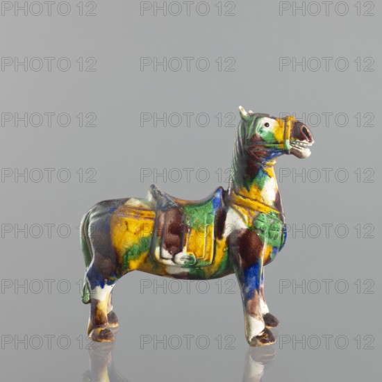Mule, Chinese, Qing dynasty 1644–1911, 18th century. Colored porcelain animal figurine sculpture of a mule. Height 19.1 cm; width 19.4 cm., A small ceramic horse figurine stands on a reflective surface against a plain gray background; the figure is glazed in a mix of colors including yellow, green, blue, brown, white, and a touch of dark red, with a saddle-like raised area on its back and stylized features on its head and mane, showing a slightly open mouth and pronounced ears, and the glaze creates mottled patches and drips across the body and legs.