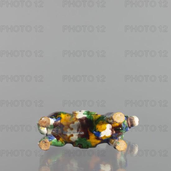 Mule; Chinese colored porcelain animal figurine sculpture, Qing dynasty 18th century, China; height 19.1 cm, width 19.4 cm., A small ceramic figurine viewed from below resting on a reflective surface, showing a mottled glaze in green, yellow, brown, blue, white, and black. The base has four rounded protrusions with exposed unglazed terracotta ends and a slightly irregular shape. The reflective surface creates a faint mirrored reflection of the figurine. The background is a smooth, neutral gray.