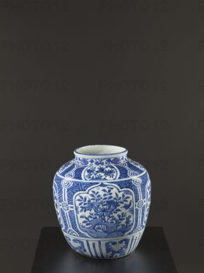 Resource pot vase, maker, Chinese blue-and-white porcelain vessel with floral decorations and flower tendril motif; decoration in a wide band with four reserves depicting rocks and flowering shrubs; Qing dynasty 1644–1911, 17th–18th century; height 35 cm; provenance unknown., A ceramic jar sits on a dark surface against a dark gray background, showing a rounded shape with a short neck and open mouth; the jar is decorated in blue and white with several panels of floral and foliage motifs, stylized borders, and repeating geometric patterns, and the visible colors include shades of blue, white, and the dark gray of the background and surface.