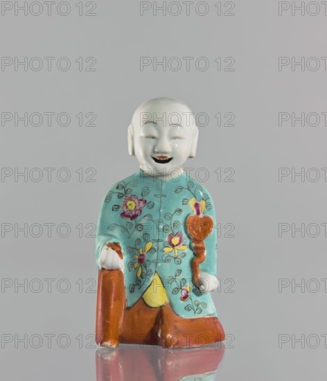 Boy with ruyi — incense burner; maker, Chinese; colored porcelain; ruyi scepter of prosperity iconography; incense burner depicting a boy/child; 19th century, China; sculpture; height 15 cm., A small figurine of a smiling person with a smooth white head and facial features, wearing a turquoise garment decorated with floral patterns in pink, yellow, and green, holding a brown staff and a rounded brown object, with brown lower garments and a yellow triangular detail at the front, all set against a plain light gray background and reflected faintly on the surface beneath.