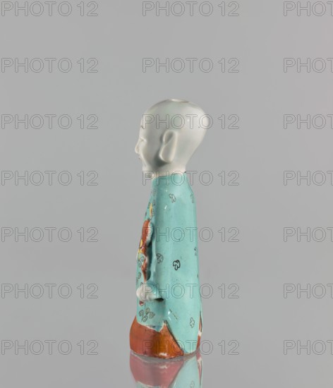 Boy with ruyi — incense burner; creator, Chinese; colored porcelain; Ming; 19th century; subject: boy holding a ruyi scepter iconography of prosperity; function: incense burner; material: porcelain; sculpture; height 15 cm., A ceramic figure shown in profile standing on a reflective surface, with a smooth pale head and facial features molded in low relief, a long teal robe decorated with small floral motifs and a painted design along the front, and a reddish-brown base; visible colors include pale white, teal, reddish-brown, and small accents of darker and lighter tones.