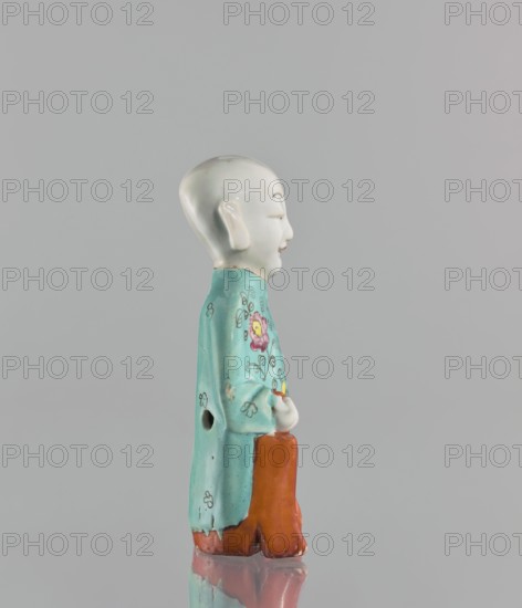 Boy with ruyi — incense burner, Chinese, 19th century, colored porcelain, Ming style, ruyi scepter of prosperity iconography, depicting a boy/child, sculpture, height 15 cm., A ceramic figurine shown in profile on a gray background, featuring a glossy white head with simple facial features and ears, a turquoise robe decorated with floral motifs and small dark accent marks, and an orange lower garment; the figure stands on a reflective surface that mirrors parts of the base, and visible colors include white, turquoise, pink, yellow, orange, dark gray, and the neutral gray of the backdrop.