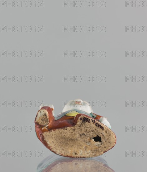 Boy with ruyi — incense burner., Chinese; colored porcelain; Ming; 19th century; subject: boy holding a ruyi scepter iconography: ruyi scepter of prosperity, incense burner; sculpture; height 15 cm., A small ceramic figurine viewed from below resting on a reflective surface, showing a worn, unglazed underside in beige and light brown with a visible hole and some dark spots; the glazed upper portions visible around the edge show glossy colors including red, white, yellow, green, and blue, with small areas of brownish wear.