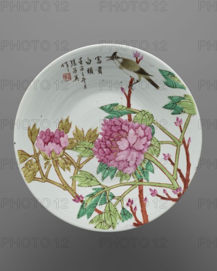 Platter, Chinese, huaniao style, 18th/19th century, China; porcelain platter decorated with birds, peonies and other flowers in colored enamel; vessel; height 4.6 cm, diameter 29.8 cm., A circular ceramic plate with a white background, decorated with a painted scene of a bird perched on a branch above a group of large blooming flowers and foliage; the bird is rendered in shades of olive green, brown, black, white, and a touch of yellow, and the branches are brown with buds in pink and red tones. The flowers are mainly pink and magenta with subtle yellow accents at their centers, surrounded by leaves in several shades of green and olive. The overall palette includes white, green, pink, magenta, brown, olive, black, and yellow, and there is a cluster of dark-colored calligraphic marks near the rim that are ignored as text. The composition fills most of the plate, leaving a band of white space near the rim, and the plate sits against a neutral gray background.