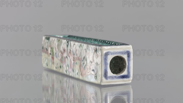 Cricket Cage, Chinese, Qing dynasty 1644–1911, colored porcelain vessel depicting animals and a cricket-fighting scene; 1st half of the 19th century; height 17.9 cm, width 4.6 cm., A rectangular ceramic object lies on a reflective surface, showing a long side with a textured, multicolored glaze and an end face with a circular opening. The surface colors visible include pale white or cream as a base, with muted green, soft pink, light blue, and touches of darker green and bluish-purple around the opening, and the interior of the opening appears dark green. The long top edge has a glossy green ribbed texture, and the long side displays an irregular pattern of raised dots and swirled glaze in the muted colors. The end face has a framed border of bluish-purple surrounding the circular opening, and the object and its colors are mirrored faintly on the shiny surface beneath. The background is a uniform light gray.