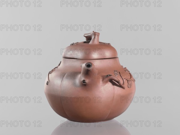 Teapot with a peach-shaped lid, maker, peaches iconography; Chinese art; teapot in Yixing style with handle and spout shaped like tree branches from which flowering branches grow around the vessel; red stoneware; 18th century; China; vessel; height 11.8 cm, width 17.5 cm, A rounded ceramic teapot in a reddish-brown color sits against a pale gray background, showing a bulbous body with a curved spout and a short handle-like protrusion on one side, a fitted lid topped by a sculpted knob, and several raised decorative elements resembling small blossoms or vines in a darker brown shade; the surface shows subtle variations in tone with areas of lighter and darker reddish-brown and a soft sheen.