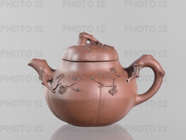 Teapot with a peach-shaped lid, maker, peaches iconography; Chinese art; teapot in Yixing style with handle and spout shaped like tree branches from which flowering branches grow surrounding the vessel; red stoneware; 18th century, China; vessel; height 11.8 cm, width 17.5 cm, A reddish-brown ceramic teapot with a rounded body, curved spout, and a looped handle sits against a pale gray background; the surface features raised branch and blossom decorations that wrap around the body and lid, and the lid has a small sculpted branch-like knob, with subtle variations of darker and lighter reddish-brown tones and small areas of darker patina.